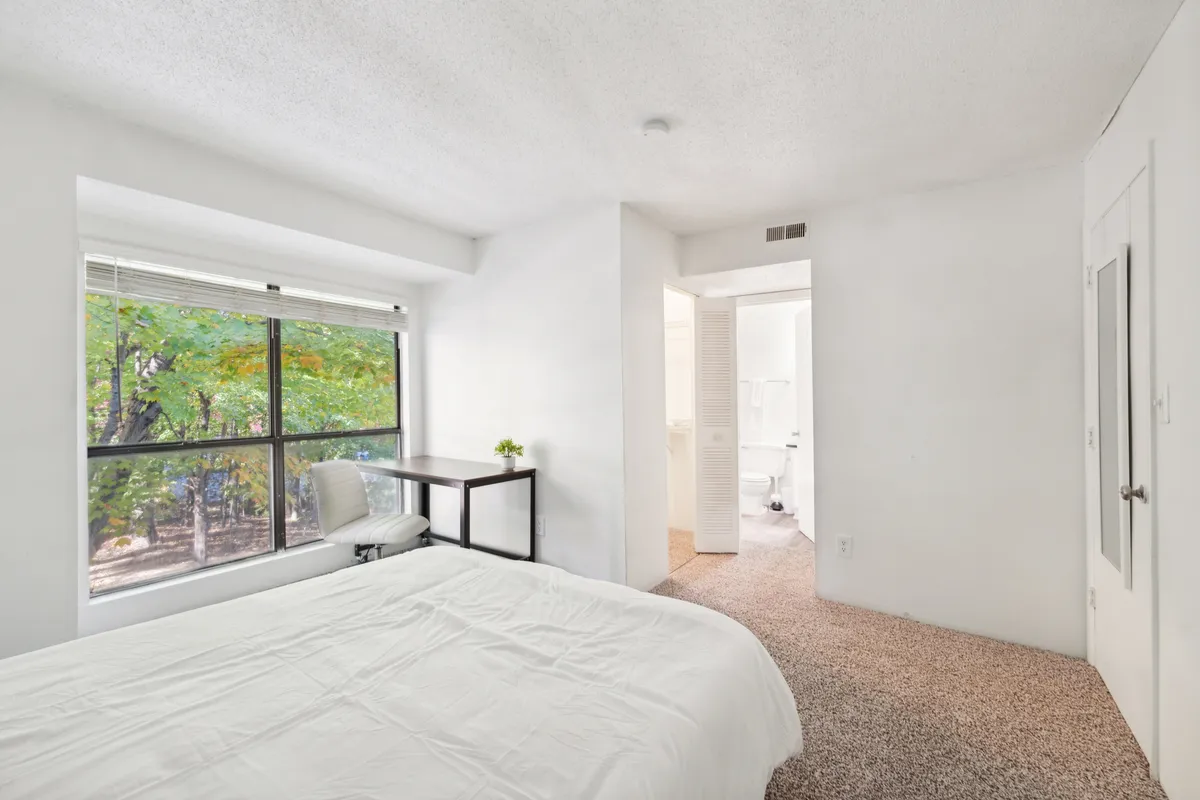 Another inviting bedroom scene, this image showcases a bed with plush white linens, a window streaming in gentle daylight, and a functional desk and chair. The room exudes comfort and tranquility, with furnishings that make it ideal for reading, working, or simply relaxing.