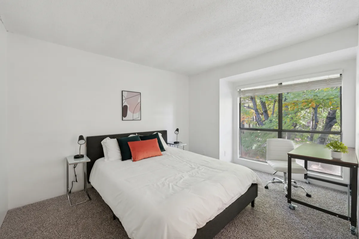 This image shows a bright and peaceful bedroom featuring a bed with crisp white bedding and accent pillows. A large window lets in natural light and offers a view of greenery, while a desk and chair make the space versatile for both work and relaxation. The decor is understated and stylish, contributing to a restful ambiance.