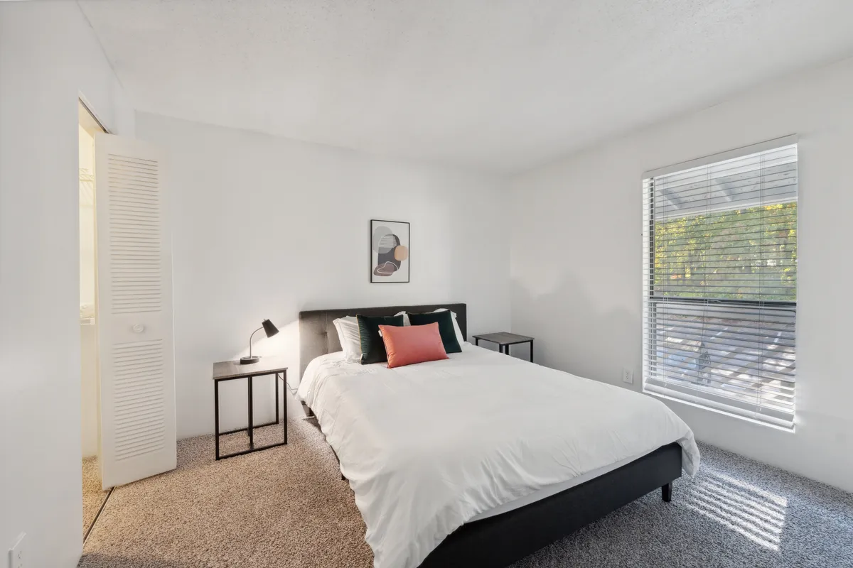 The image showcases a serene and inviting bedroom featuring a plush bed with crisp white bedding and stylish accent pillows, complemented by modern framed artwork on the wall and a nightstand softly illuminated by a lamp. The space exudes tranquility and comfort, ideal for relaxation and restful nights.